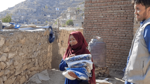 A woman in a red headscarf holds folded clothing outdoors, with hillside homes and a brick wall in the background