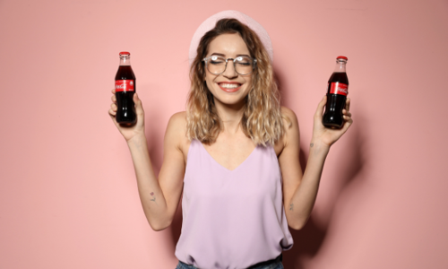 Smiling young woman in a pink top and hat holding two glass bottles of Coca-Cola in front of a pink background