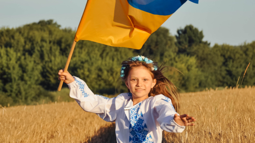 Child wearing traditional embroidered clothing waves a blue-and-yellow flag in a wheat field